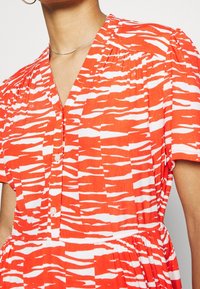 Czerwona i biała sukienka w wzory z krótkimi rękawami, dekoltem w serek, zapięciem na guziki i marszczeniami na ramionach, wykonana z lekkiego materiału.