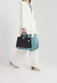 Donna che indossa un blazer bianco, un vestito teal, pantaloni bianchi larghi e tacchi beige, mentre porta una borsa nera strutturata con dettagli dorati.