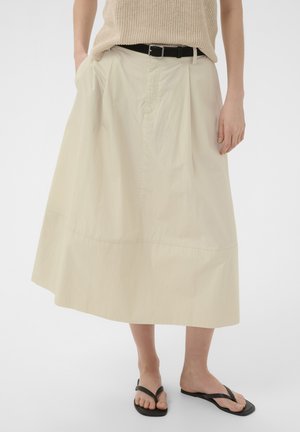 Kobieta ubrana w beżową spódnicę midi z czarnym paskiem, beżowy dziergany top i czarne sandały na palce, stoi ze swobodnie opuszczonymi rękami.