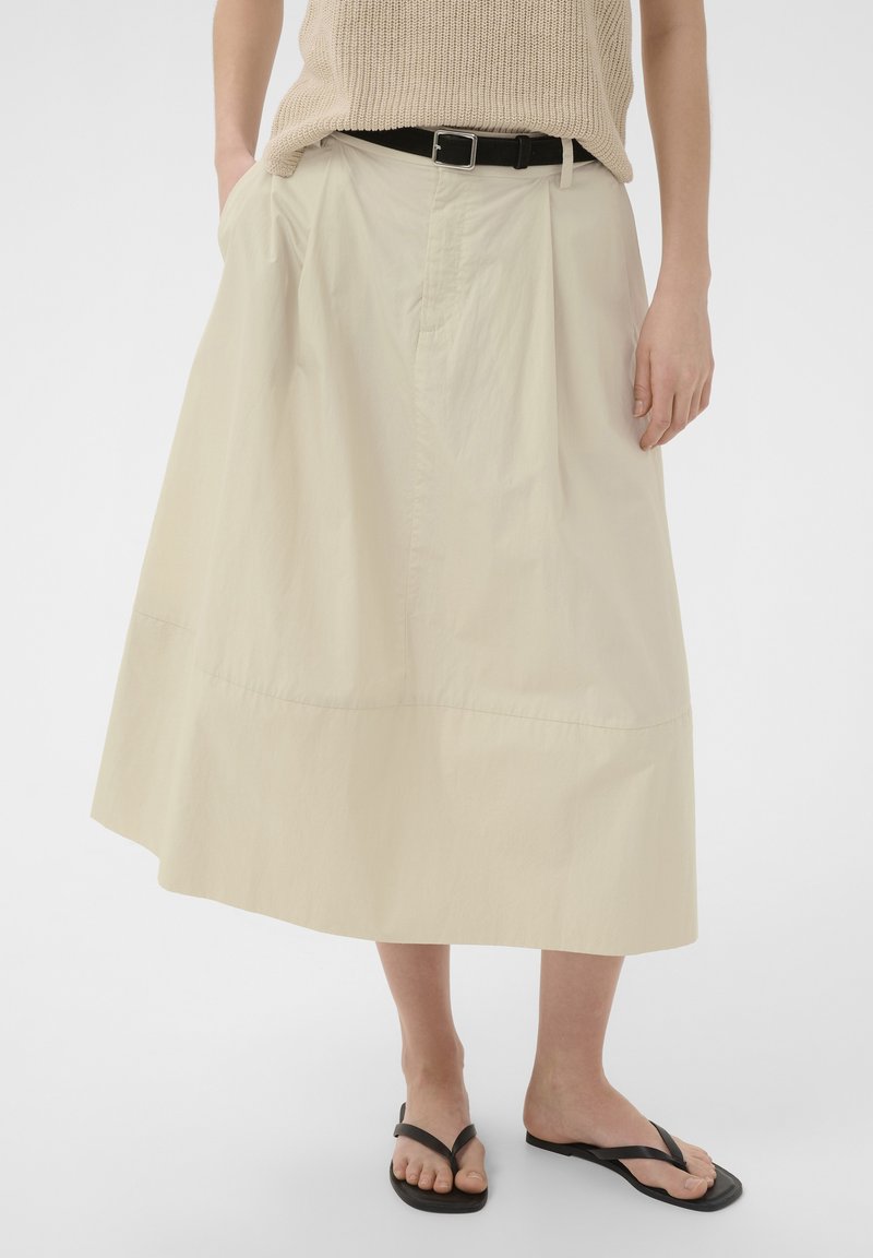 Femme portant une jupe midi beige avec une ceinture noire, un haut en maille beige et des sandales tongs noires, debout les mains détendues.