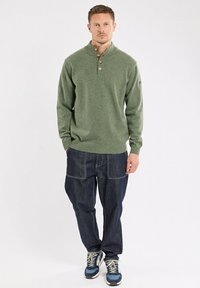 Pull en tricot vert avec un col, trois boutons et une finition côtelée, associé à un pantalon en jean foncé ample et des baskets bleues.