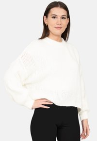 Jonge vrouw met stijlbruin haar draagt gouden oorringen, een witte grofgebreide trui en zwarte broek, poserend met een hand op haar heup.