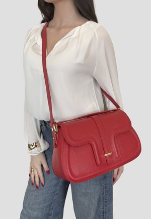 Donna che indossa una camicetta bianca e jeans blu, che tiene una borsa a tracolla rossa in pelle testurizzata con dettagli in oro.