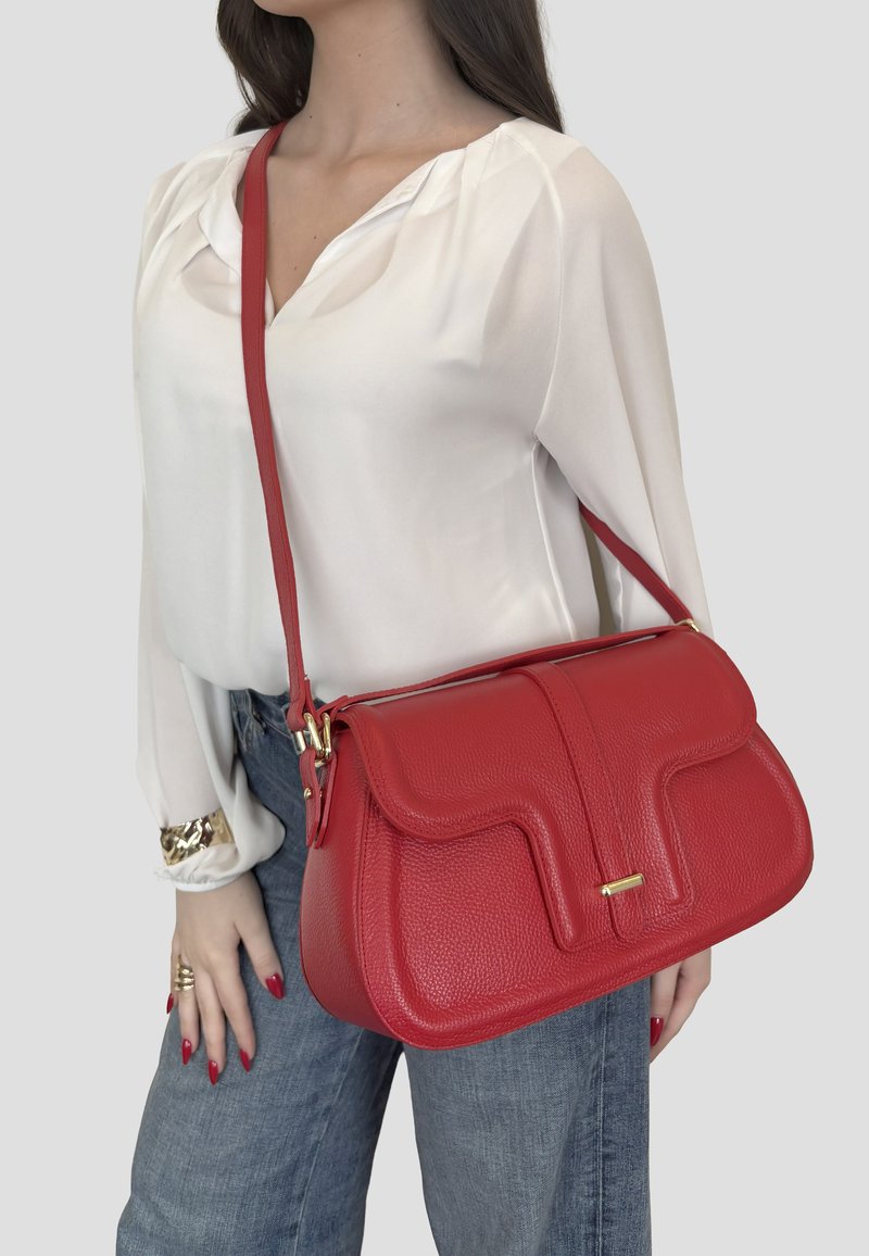 Donna che indossa una camicetta bianca e jeans blu, che tiene una borsa a tracolla rossa in pelle testurizzata con dettagli in oro.