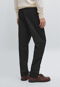 Pantaloni neri con una vestibilità affusolata, realizzati in tessuto morbido, dotati di tasche laterali e una texture sottile, abbinati a scarpe marroni con lacci.