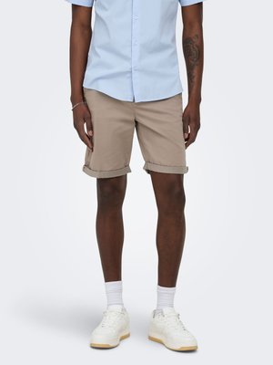 Homme portant un short beige retroussé, une chemise bleu clair boutonnée, des chaussettes blanches et des baskets blanches, debout devant un fond uni.