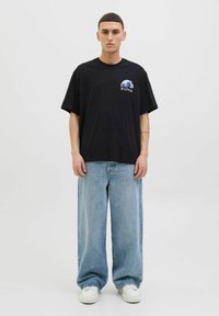 Camiseta negra de corte holgado con un gráfico de olas y texto en japonés en el frente, combinada con jeans de pierna ancha en azul claro y zapatillas blancas.
