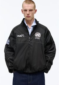 Mladý muž s krátkými vlasy, oblečený v černé sportovní bundě se zipem označené "USA 91" a "SPORT", stojí s rukama v kapsách na bílém pozadí.