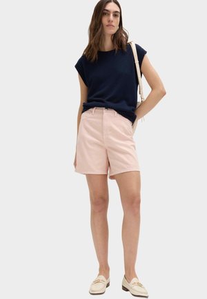 Frau trägt marineblaues ärmelloses Oberteil, hellrosa knielange Shorts, beige Mokassins und eine weiße Umhängetasche, steht mit einer Hand in der Hüfte.