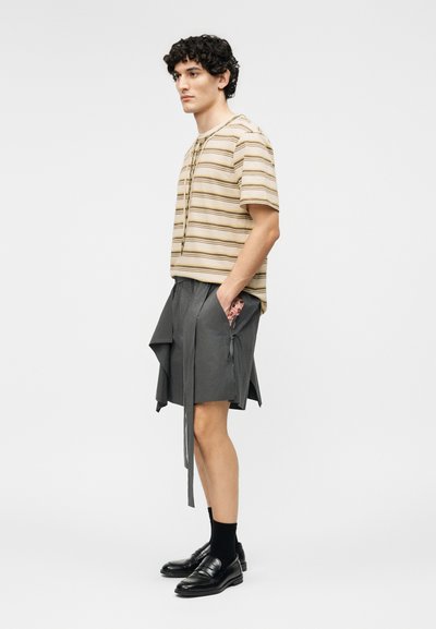 Jeune homme portant une chemise à manches courtes à capuche beige rayée, un short gris mi-cuisses avec des sangles pendantes, des chaussettes noires et des mocassins noirs.