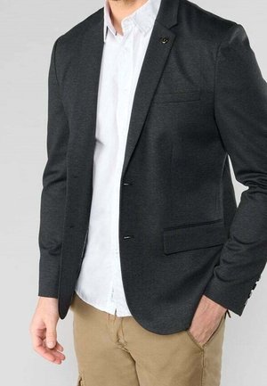 Homme portant un blazer gris foncé sur une chemise blanche boutonnée et un pantalon beige, une main dans la poche, sur un fond uni.