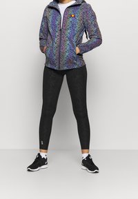 Färgglad mönstrad zip-jacka med huva och två sidofickor, kombinerad med svarta tajta leggings och svarta sportskor.