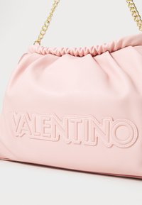 Rozā ādas rokassomiņa ar saliektu dizainu, zelta ķēdes siksnu un iegrieztu "VALENTINO" logo priekšpusē. Gluda tekstūra.