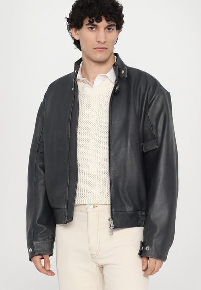 Veste en cuir noir avec fermeture éclair à l'avant, col à boutons-pression et détails élastiques sur les côtés. Associée à un pull en tricot crème et un pantalon clair.
