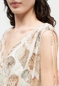 Γυναίκα φορούν αμάνικο φόρεμα με V-λαιμόκοψη και σχέδιο με κοχύλια, με λεπτές τιράντες στους ώμους, που δείχνει το λαιμό και τους άνω ώμους της.