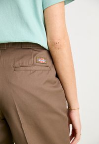 Pantalons marron avec une texture lisse et une poche arrière, arborant un écusson logo Dickies rouge, jaune et bleu. Chemise teal visible au-dessus.