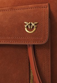 Braune Wildledertasche mit goldfarbenem Metalllogo, das zwei Vögel zeigt, weiche Textur und sichtbare Nähte. Reißverschlussdetails an der Seite.