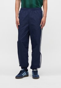 Παντελόνι φόρμας ναυτικού μπλε από ελαφρύ ύφασμα, με λευκές ρίγες στο πλάι και ελαστική μέση. Συνδυάζεται με μπλε αθλητικά παπούτσια.