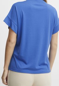Donna che indossa una camicia blu a maniche corte con risvolti e pantaloni beige, vista da dietro su uno sfondo semplice.
