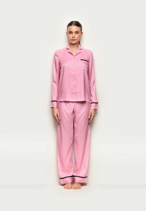 Frau steht barfuß und trägt glänzende rosa Satin-Pyjamas mit schwarzem Besatz an Bündchen, Tasche und Hosenenden vor hellem, einfarbigem Hintergrund.