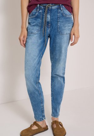 Persona che indossa pantaloni jogger in denim azzurro chiaro con tasche frontali e vita con coulisse, abbinati a scarpe slip-on in camoscio marrone.