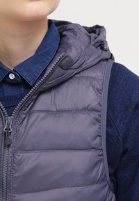 Gepolsterte graue Weste mit Kapuze, Reißverschluss vorne und subtiler Schattierung. Kombiniert mit einem dunkelblauen Strickhemd mit Kragen und Knöpfen.