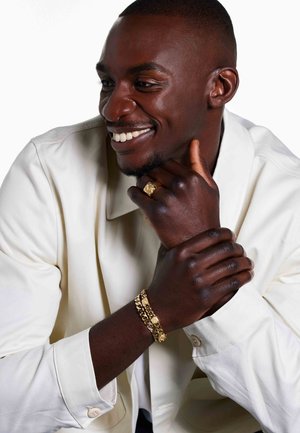 Bague en or avec un détail texturé et un bracelet en or massif sur la main d'un homme à la peau foncée, portant une chemise beige clair, souriant.