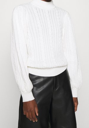 Personne portant un pull en maille torsadée blanc à col haut et manches longues, associé à un pantalon en cuir noir, debout devant un fond uni.