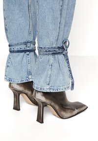 Zapatos de tacón alto de piel marrón con puntera afilada, combinados con pantalones de mezclilla azul claro con dobladillo fruncido y lazos.