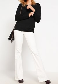 Schwarzes Langarmoberteil, weiße Schlaghosen und dunkle Absatzschuhe. Das Modell hält eine Fransen-Crossbody-Tasche. Glattes Material mit lässiger Passform.
