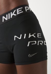 Czarne szorty Nike Pro z szerokim pasem, na którym znajduje się biały napis "NIKE PRO" oraz białe akcenty logo na nogawce. Gładki, elastyczny materiał.