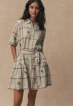 Frau mit dunklen, lockigen Haaren trägt ein beiges Hemdkleid mit braunen, gestickten Blumenmustern, kurzen Ärmeln und einem gebundenen Taillengürtel.