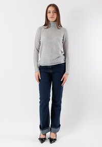 Maglione grigio a collo alto, stile aderente, indossato con jeans in denim blu scuro, risvoltati in fondo. Tacchi neri a punta completano l'outfit.
