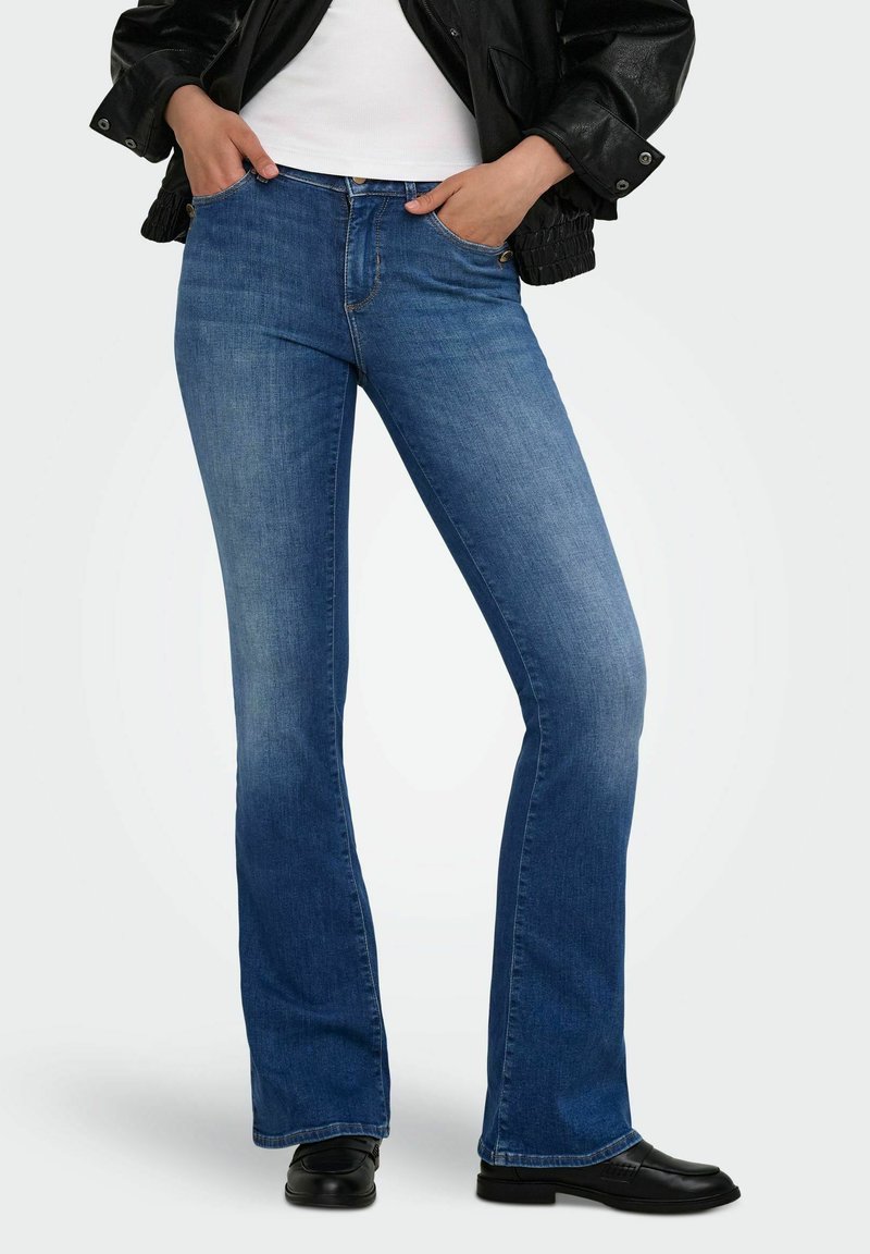Femme portant un jean bootcut bleu, des mocassins en cuir noir, un haut blanc, et une veste noire avec les mains dans les poches.