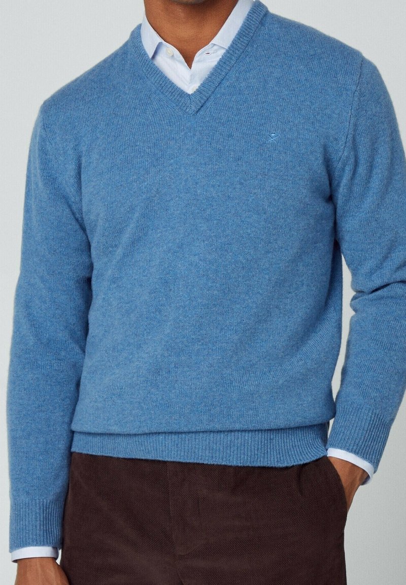 Pull en laine bleu clair avec col en V, poignets et ourlet côtelés, présentant un petit logo sur la poitrine. Assorti à un pantalon marron foncé.