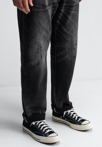 Beine in schwarzen, ausgewaschenen Jeans mit aufgerolltem Saum und schwarzen Low-Top-Sneakern mit weißen Schnürsenkeln und Sohle auf einem weißen Boden.