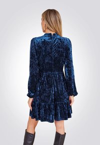 Blauwe fluwelen jurk met lange mouwen, gerimpelde zoom en hoge halslijn. Kenmerkt zich door textuurcontrast en een aansluitende taille.