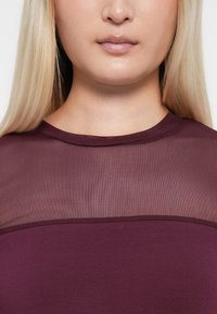 Burgundy topp med en genomskinlig meshpanel vid halsringningen, med rund krage och slät, åtsittande tygstruktur.