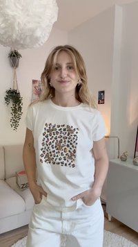 White short-sleeve t-shirt featuring a leopard print pattern in brown and black. Model wears white pants, standing with hands in pockets.