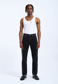 Jeans de mezclilla negra con un corte recto, que presentan una textura suave y costuras visibles. Combinados con una camiseta sin mangas blanca y collares superpuestos.