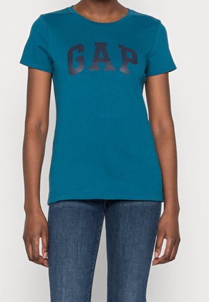 Osoba nosząca zielononiebieski krótki rękaw T-shirt marki GAP i niebieskie dżinsy, stojąca na jednolitym białym tle.