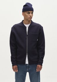 Mornarska zip-up jakna iz teksturiranega blaga, z izdignjenim ovratnikom, sprednjimi žepti in ozkim krojem. Nošena čez belo srajco in modre kavbojke.