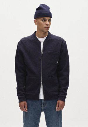 Tumesinine tõmblukuga jakk, mis on valmistatud tekstuuriga kangast, seisva kraega, ees olevate taskutega ja kitsastes lõigetega. Kandmine valge särgi ja siniste teksade peal.