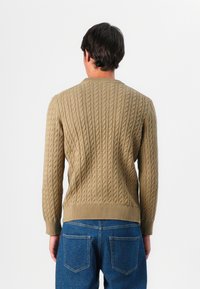 Pull en tricot beige avec un col rond et des bords côtelés, présentant un motif texturé. Associé à un jean en denim bleu.