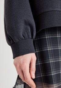 Tmavošedý sveter s rebrovanými manžetami a objemnými rukávmi, skombinovaný s čierno-bielou kockovanou sukňou. Ruka na obrázku je uvoľnená.