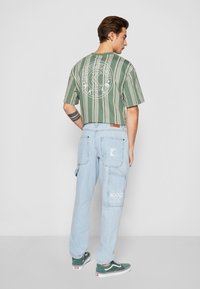 Chemise à rayures vertes à manches courtes avec un graphique circulaire blanc au dos, associée à un pantalon cargo bleu clair et des baskets vertes.