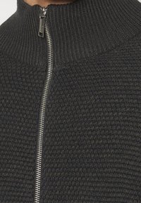 Stickad tröja i mörkgrått med hög krage och dragkedja fram. Har ett texturerat tyg med ribbade detaljer vid kragen.