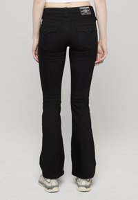 Zwarte uitlopende spijkerbroek met achterzakken voorzien van decoratieve stiksels en metalen accenten. Draagt grijs-witte sneakers, minimale zichtbare textuur.