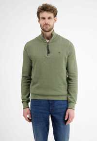 Grüner Pullover aus strukturiertem Strickmaterial mit einem Reißverschlusskragen, kombiniert mit blauen Jeans. Verfügt über ein kleines Logo auf der linken Brust.