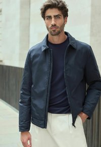 Jachetă bomber bleumarin cu textură netedă, închidere cu fermoar și design cu guler, purtată peste o cămașă închisă la culoare și pantaloni de o nuanță deschisă.